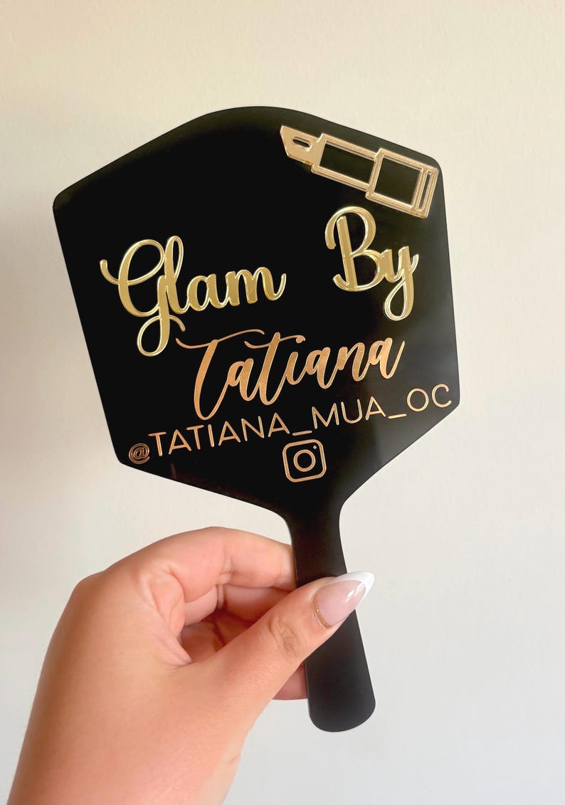 Może przedstawiać: Czarne lusterko ręczne o sześciokątnym kształcie ze złotym napisem Glam By Tatiana, uchwytem @TATIANA_MUA_OC, ikoną Instagrama oraz złotą grafiką szminki umieszczoną w prawym g&oacute;rnym rogu powierzchni lustra.