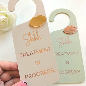 May include: Two door hangers, one beige and one sage green, each featuring a gold or rose gold mirrored lip icon at the top. The text "Shhhh TREATMENT IN PROGRESS..." is printed in a metallic script and block font on the front of each hanger.