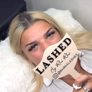 May include: A beige, fan-shaped handheld sign with a handle, featuring the black text "LASHED By Rhi Rhi" and an Instagram icon followed by "@RHIRHI.LASHES". The sign is held in front of the face, partially obscuring the nose and mouth area.