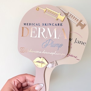 May include: Two handheld paddle mirrors in dusty rose and beige. The front mirror features a gold syringe icon, the text MEDICAL SKINCARE DERMA Plump, an Instagram icon, and the handle @skincare.dermaplumpx with a gold metallic lip emblem.
