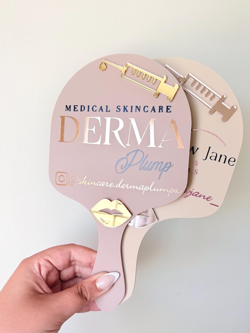 May include: Two handheld paddle mirrors in dusty rose and beige. The front mirror features a gold syringe icon, the text MEDICAL SKINCARE DERMA Plump, an Instagram icon, and the handle @skincare.dermaplumpx with a gold metallic lip emblem.