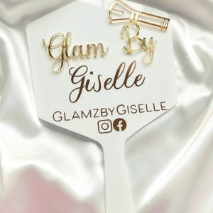 Może przedstawiać: Białe lusterko ręczne ze złotym napisem Glam By Giselle, GLAMZBYGISELLE oraz ikonami medi&oacute;w społecznościowych. Złota spinka do włos&oacute;w jest przymocowana do prawego g&oacute;rnego rogu powierzchni lustra, kt&oacute;re ma gładki, płaski, sześciokątny kształt.