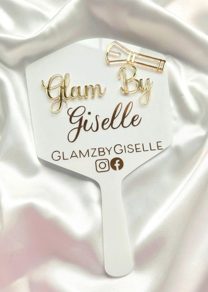 Może przedstawiać: Białe lusterko ręczne ze złotym napisem Glam By Giselle, GLAMZBYGISELLE oraz ikonami medi&oacute;w społecznościowych. Złota spinka do włos&oacute;w jest przymocowana do prawego g&oacute;rnego rogu powierzchni lustra, kt&oacute;re ma gładki, płaski, sześciokątny kształt.