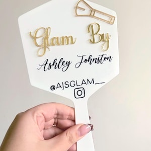 Może przedstawiać: Białe lusterko ręczne ze złotymi literami 3D z napisem Glam By. Poniżej nadrukowano czarny tekst Ashley Johnston, a następnie nazwę użytkownika Instagram @AJSGLAM__ z ikoną. Dołączona jest złota ikona pędzla do makijażu.