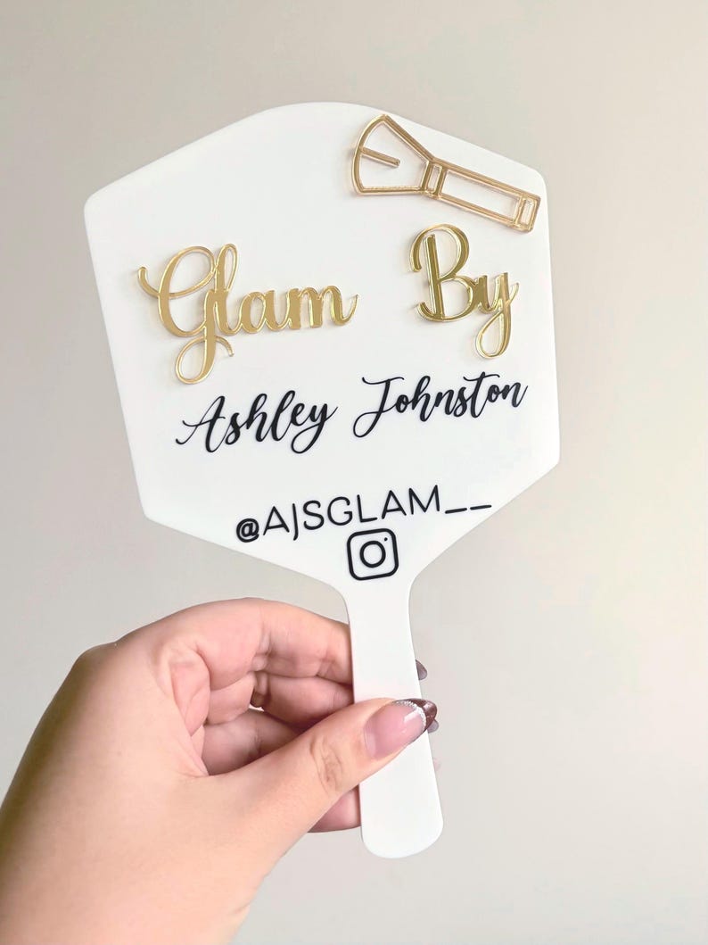 Może przedstawiać: Białe lusterko ręczne ze złotymi literami 3D z napisem Glam By. Poniżej nadrukowano czarny tekst Ashley Johnston, a następnie nazwę użytkownika Instagram @AJSGLAM__ z ikoną. Dołączona jest złota ikona pędzla do makijażu.