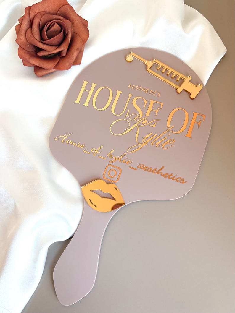 May include: A mauve handheld mirror featuring gold-toned accents including a syringe icon, a lip icon, and an Instagram logo. The text reads HOUSE OF AESTHETICS, Kylie, and house_of_kylie_aesthetics in a mix of block and script fonts.