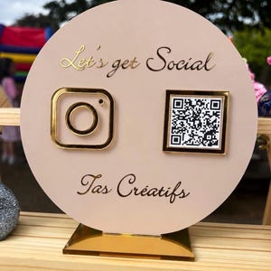 May include: A circular, blush-toned sign with gold detailing. It displays an Instagram logo, a QR code, and the phrases "Let's get Social" and "Tas Créatifs" in gold script. The sign is mounted on a gold base.