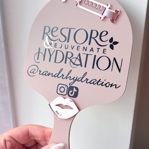 May include: A pale pink handheld sign featuring a rose gold acrylic syringe icon at the top and a rose gold lip icon at the bottom. The text reads RESTORE & REJUVENATE HYDRATION @randrhydration with Instagram and TikTok social media icons.
