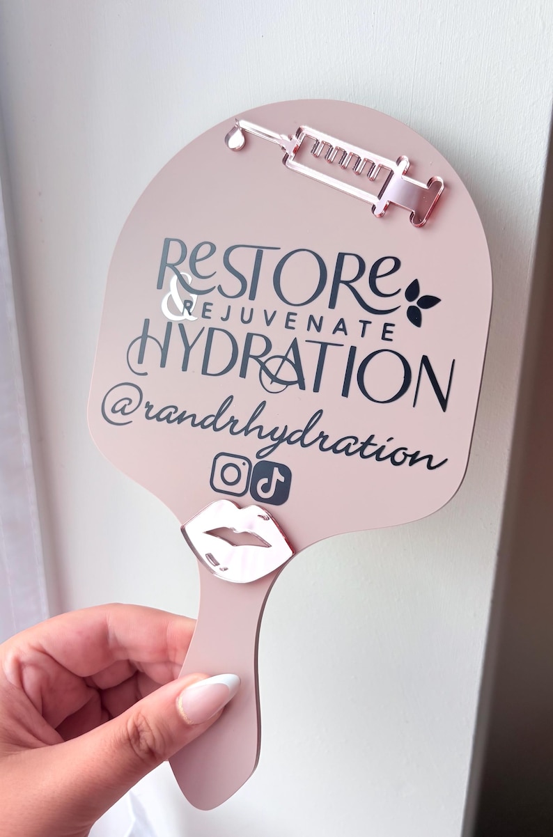 May include: A pale pink handheld sign featuring a rose gold acrylic syringe icon at the top and a rose gold lip icon at the bottom. The text reads RESTORE & REJUVENATE HYDRATION @randrhydration with Instagram and TikTok social media icons.
