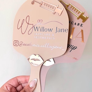 May include: Two handheld paddle mirrors in light beige and dusty rose. The beige mirror features a rose gold syringe icon, a silver lip icon, and the text Willow Jane Skincare & Aesthetics with an Instagram handle. The rose mirror shows a gold syringe.