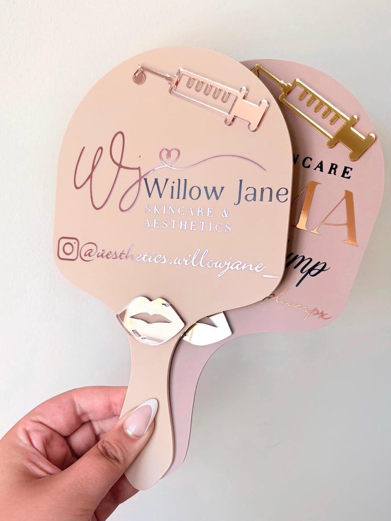 May include: Two handheld paddle mirrors in light beige and dusty rose. The beige mirror features a rose gold syringe icon, a silver lip icon, and the text Willow Jane Skincare & Aesthetics with an Instagram handle. The rose mirror shows a gold syringe.