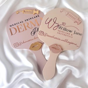 May include: Two light pink handheld mirrors with handles. The left mirror features the text MEDICAL SKINCARE DERMA and an Instagram handle, with a gold syringe and lip icon. The right mirror displays Wj Willow Jane SKINCARE & AESTHETICS.