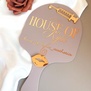 May include: A mauve handheld mirror featuring gold-toned accents including a syringe icon, a lip icon, and an Instagram logo. The text reads HOUSE OF AESTHETICS, Kylie, and house_of_kylie_aesthetics in a mix of block and script fonts.