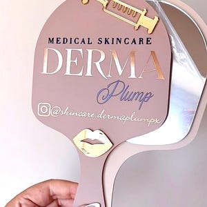 May include: A dusty rose handheld mirror featuring a gold syringe icon at the top and a gold lip icon on the handle. The text reads MEDICAL SKINCARE DERMA Plump with an Instagram icon and the handle @skincare.dermaplumpx in white lettering.