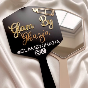 Może przedstawiać: Czarne lusterko ręczne ze złotą ikoną szminki i złotym napisem Glam By Ghazia. Pod tekstem znajduje się biały uchwyt @GLAMBYGHAZIA z ikonami Instagrama i TikToka. Drugie beżowe lusterko ręczne jest częściowo widoczne za czarnym lusterkiem.