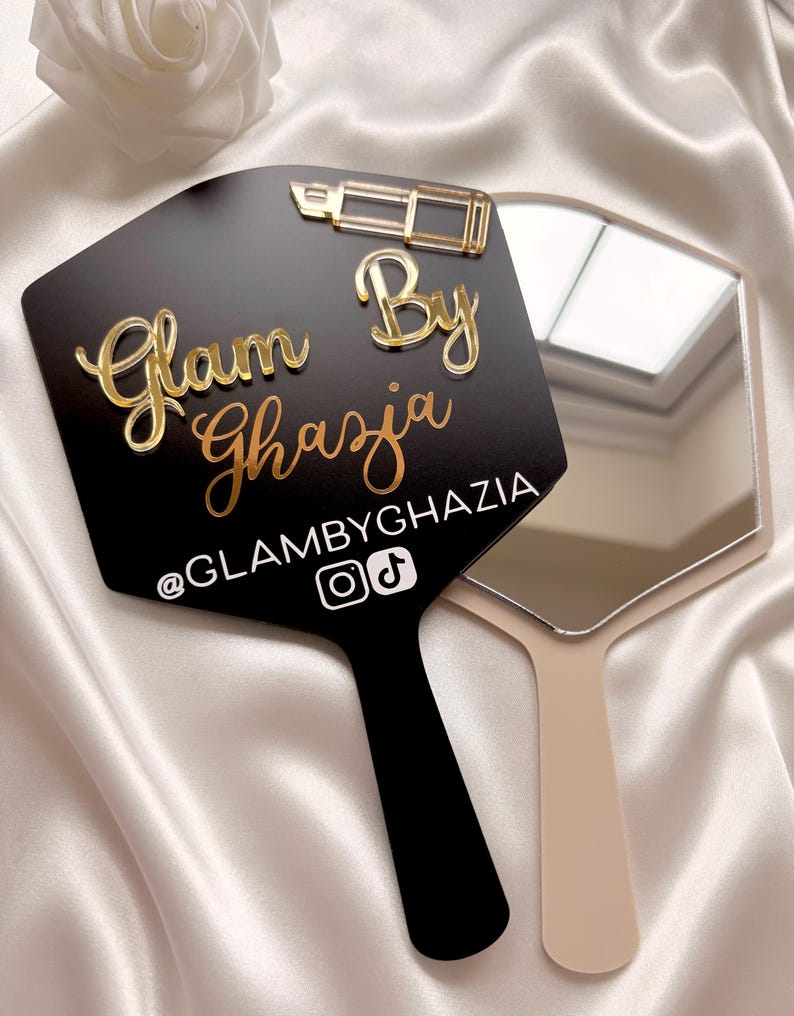 Może przedstawiać: Czarne lusterko ręczne ze złotą ikoną szminki i złotym napisem Glam By Ghazia. Pod tekstem znajduje się biały uchwyt @GLAMBYGHAZIA z ikonami Instagrama i TikToka. Drugie beżowe lusterko ręczne jest częściowo widoczne za czarnym lusterkiem.