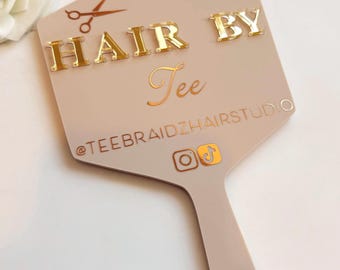Może przedstawiać: Beżowe lusterko ręczne o sześciokątnym kształcie ze złotymi literami 3D z napisem HAIR BY Tee. Poniżej tekstu znajduje się nazwa użytkownika @TEEBRAIDZHAIRSTUDIO oraz ikony mediów społecznościowych Instagram i TikTok. Widoczne są małe nożyczki.