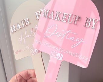 May include: Two arch-shaped acrylic handheld signs with handles. The front sign is pink with silver 3D text reading MAKEUP BY DESTINY, white script text, and social media icons. The back sign is beige with silver text reading HAIR BY HALLIE.
