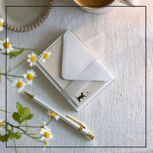 May include: A stationery set with a white pen with gold detailing, cream notecards, and a white envelope. A black cat silhouette is printed on the notecard. Daisies are in the background, on a white tablecloth.