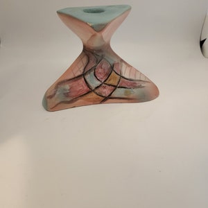 May include: A ceramic candle holder with an abstract design in shades of pink, blue, and brown. The holder has a triangular base and a unique, asymmetrical shape.