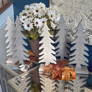 Puede incluir: Cinco árboles decorativos de madera en gris y blanco con una maceta marrón de flores blancas en el fondo. Los árboles están sobre una superficie de espejo con hojas de otoño y una pequeña figura de pájaro.