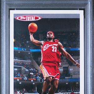 Op de afbeelding: Een 2004 Topps Total basketbal trading card met LeBron James in een rood en goud Cleveland Cavaliers uniform, een basketbal vasthoudend en in de lucht springend. De kaart is genummerd #5 en beoordeeld als NM-MT 8 door PSA.