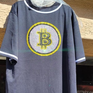 May include: A dark blue baseball jersey with a large, white, and yellow rhinestone Bitcoin logo on the back.
