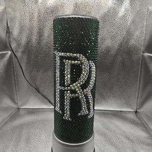 May include: A green tumbler with a white rhinestone "R" design. The tumbler has a silver base.
