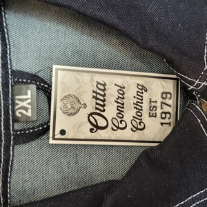 May include: Close-up of a dark blue denim jacket with white stitching. A tag reads "Outta Control Clothing EST 1979" and indicates a size of 2XL. The jacket has a button closure and a classic design.