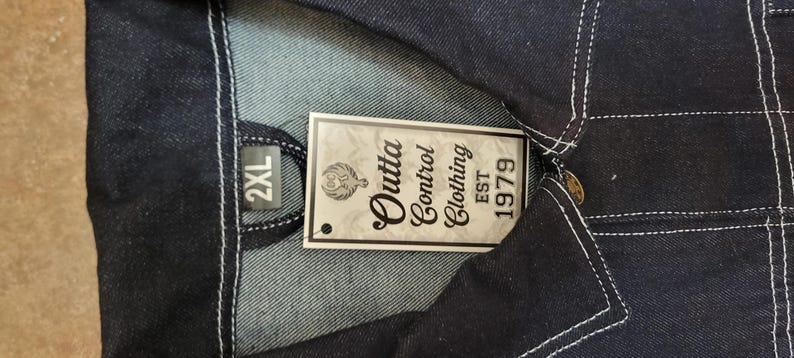 May include: Close-up of a dark blue denim jacket with white stitching. A tag reads "Outta Control Clothing EST 1979" and indicates a size of 2XL. The jacket has a button closure and a classic design.
