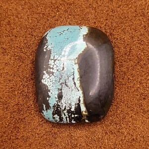 May include: A polished, oval-shaped turquoise stone with a dark, almost black, side. The turquoise section features a light blue color with white matrix patterns. The stone is set against a brown, textured background.