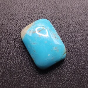 May include: A polished, rectangular turquoise gemstone with a rounded top. The stone features a vibrant, light blue color with brown and white inclusions. The gemstone is smooth and reflective, showcasing natural variations.