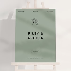 Puede incluir: Un letrero de bienvenida de boda verde salvia sobre un caballete de madera. El letrero presenta los nombres "Riley & Archer" en negrita, con la fecha "09.16.26" y las palabras "Bienvenida a nuestra boda" en la parte superior y "Nos alegra mucho que estés aquí" en la parte inferior.