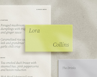 May include: A pale green name card with the name "Lora" printed in black. The word "Collins" is printed in black on the bottom of the card. The card is placed on a white menu with the words "Dinner Menu" at the top.