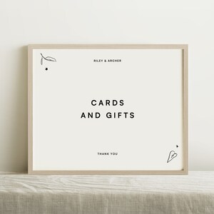 Puede incluir: Un cartel blanco con marco de madera, con el texto "Riley & Archer" en la parte superior, "Tarjetas y Regalos" en el centro y "Gracias" en la parte inferior. Hay dibujos simples de una hoja, una estrella y un corazón en el cartel.