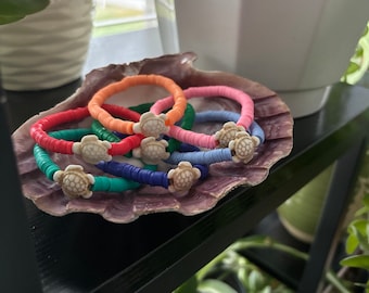 Könnte beinhalten: Ein Set aus bunten Perlenarmbändern mit einem weißen Schildkrötenanhänger an jedem. Die Armbänder sind auf einer Muschel angeordnet.