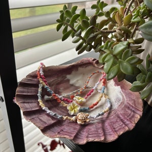 May include: Three colorful friendship bracelets with white flower and turtle charms are displayed on a pink seashell.