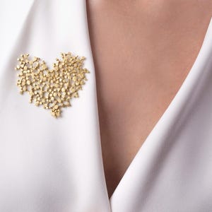 Peut inclure: Broche dorée en forme de cœur, composée de petits éléments regroupés. La broche est épinglée sur un blazer blanc. Le design est complexe et détaillé, créant un aspect texturé. La broche est un accessoire décoratif.