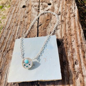 Puede incluir: Un collar de plata con un colgante circular con una piedra turquesa en el centro, rodeada de piedras verdes más pequeñas y un borde plateado texturizado. El collar se exhibe sobre un cuadrado azul claro contra una superficie de madera envejecida.