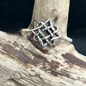 May include: A silver Celtic knot ring with a square design. The ring is displayed on a piece of driftwood.