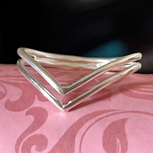 Può includere: Un braccialetto in argento a doppio strato con un design a forma di V. Il braccialetto è su uno sfondo rosa con un motivo floreale.