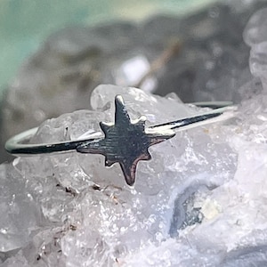 Puede incluir: Un anillo de plata con un diseño de estrella. El anillo tiene una banda delgada y un dije de estrella detallado. La estrella es de color plata mate. El anillo es una pieza de joyería delicada.