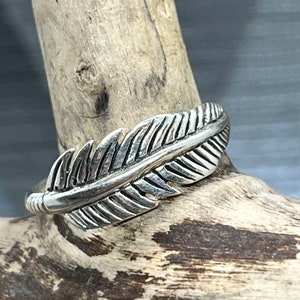 May include: A silver ring featuring a detailed feather design. The ring is displayed on a piece of driftwood.