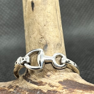 May include: A silver-colored ring featuring a horse bit design. The ring is displayed on a piece of wood, showcasing its equestrian-inspired style. The ring's design includes a central bit and connecting rings.