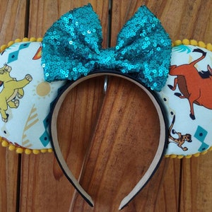 May include: A pair of mouse ears featuring characters from The Lion King. The ears have a teal sequined bow and are decorated with Simba, Timon, and Pumbaa. The edges are trimmed with yellow pom-poms, and the headband is black.