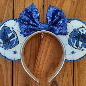 May include: Blue and white Ravenclaw themed Minnie Mouse ears with a blue sequin bow. The ears feature the Ravenclaw crest with the words "Ravenclaw" on the crest.