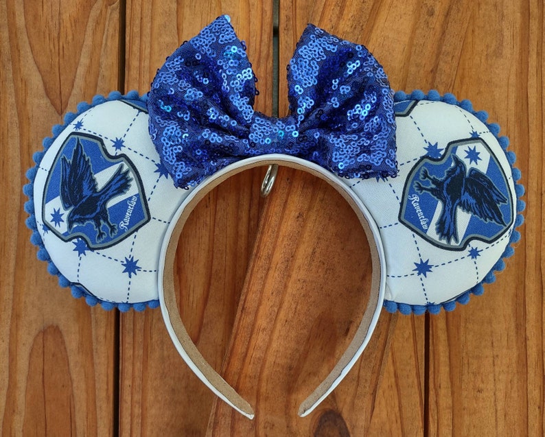 May include: Blue and white Ravenclaw themed Minnie Mouse ears with a blue sequin bow. The ears feature the Ravenclaw crest with the words "Ravenclaw" on the crest.