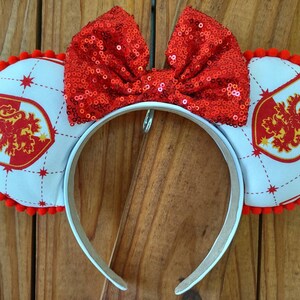 May include: White headband with red pom-poms and a red sequined bow. The headband features a white fabric with a red and gold Gryffindor crest on each side.