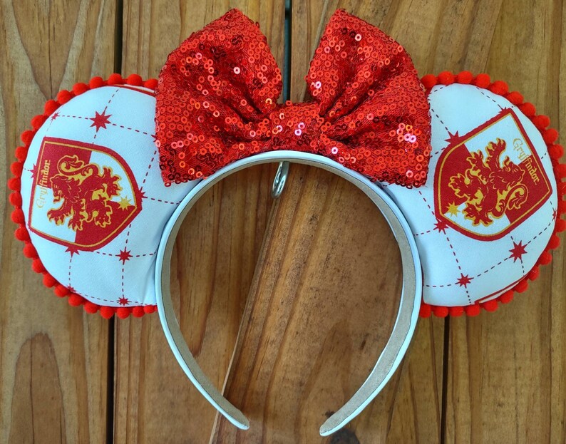 May include: White headband with red pom-poms and a red sequined bow. The headband features a white fabric with a red and gold Gryffindor crest on each side.