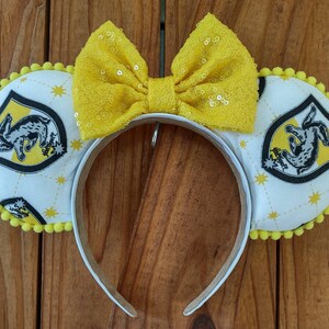 May include: Yellow and white fabric headband with a yellow bow and a white fabric patch with a black and yellow crest featuring a rabbit and the words "Hufflepuff" and "House".